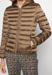 Braune, gesteppte Zip-Jacke mit Kapuze, getragen von einer Person mit einer Hand in der Tasche, kombiniert mit mehrfarbig gemusterten Hosen.