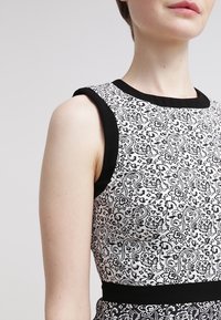 Svartvit mönstrad klänning med en åtsittande bodice, ärmlös design och svarta detaljer runt halsen och ärmarna. Har ett blommönster.