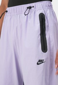 Lavandas Nike šortos ar elastīgu jostu, melna rāvējslēdzēja kabata sānos un diskrēta melna logotipa akcenta uz kājas.