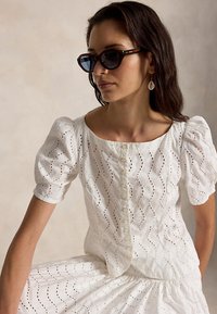 Γυναίκα που φοράει γυαλιά ηλίου από κέλυφος χελώνας, λευκό πουκάμισο με κουμπιά και φουσκωτά μανίκια από ύφασμα με τρύπες, φούστα που ταιριάζει και σκουλαρίκια με σταγόνες κοχυλιών, καθισμένη μπροστά σε μπεζ φόντο.