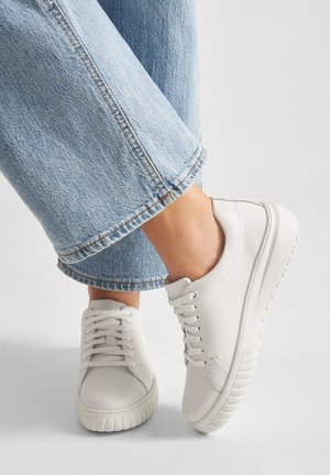 Bílé kožené tenisky s kulatou špičkou, plochými tkaničkami a gumovou podrážkou. Nošeny s světlými modrými oříznutými džínami s ohrnutými lemy.