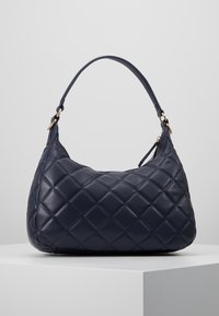 Marinblå kviltad läderväska med rundad form, med en kort axelrem och detaljer i guldton, uppvisad på en ljus pelare.