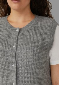 Grijs gebreid vest met een gemêleerd patroon, ronde halslijn en knopen aan de voorkant. Draagt over een wit shirt met korte mouwen.