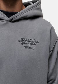 Felpa grigia con cappuccio dotata di una grande tasca frontale, cordoncino e testo ricamato in nero che recita "Good For Nothing, Established 2011."