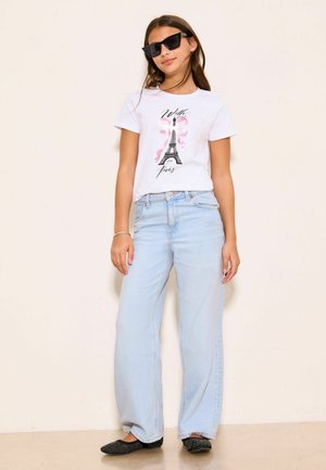 Meisje met zwarte zonnebril, wit T-shirt met Eiffeltoren-ontwerp, lichtblauwe jeans met wijde pijpen en zwarte platte schoenen met studs, staand binnen.