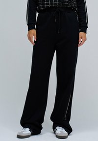 Personne portant un pantalon large noir avec une taille élastique et des baskets noir et blanc, sur fond gris uni.