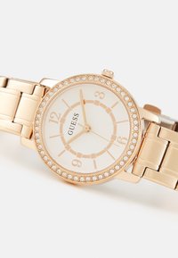 Auksinės spalvos laikrodis su apvaliu ciferblatu, turinčiu baltą dialą, auksinius valandų žymeklius ir kristalų akcentuotą rėmelį. Metalinė nuorodų apyrankė.