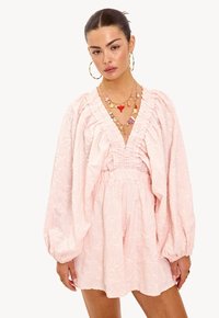 Roze jurk met lange pofmouwen, geplooide details bij de taille, een textuur met bloemmotieven en een diepe V-halslijn. Gelaagde gouden kettingen accentueren.