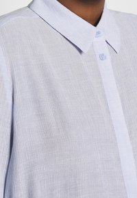 Camicia colletto azzurro fatta di tessuto texturizzato, con una placca di bottoni con bottoni blu e un motivo sottile.