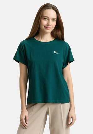 Frau trägt ein dunkelgrünes T-Shirt mit kurzen Ärmeln, einem kleinen weißen Herzlogo und beigen Hosen, steht vor einem schlichten Hintergrund.