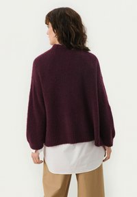 Maglione oversize di un profondo color bordeaux con maniche larghe e orlo a costine, indossato sopra una camicia bianca, realizzato in un morbido tessuto testurizzato.