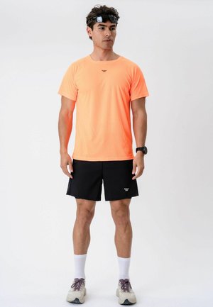 Homme portant un t-shirt orange, un short noir, des chaussettes blanches et des baskets, debout avec une lampe frontale et une montre sur fond blanc.