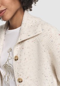 Primo piano di una persona che indossa un cardigan crema maculato con grandi bottoni e una camicia bianca con un disegno astratto dorato.
