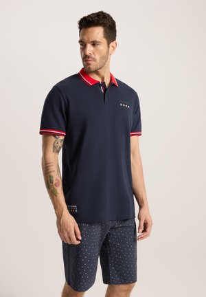 Granatowa koszulka polo z czerwonym kołnierzykiem i akcentami na rękawach, z logo i kolorowymi flagami nad dołem; zestawiona z granatowymi szortami w białe kropki.