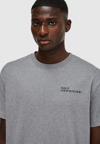 Grijze katoenen t-shirt met korte mouwen, een ronde hals en zwarte tekst met de tekst "HALO 196819721981" op de linkerborst.