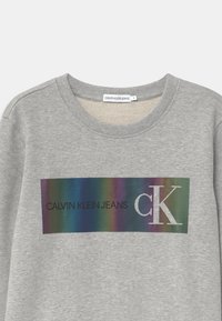 Šedá mikina s multibarevným obdélníkovým grafickým designem, na kterém je nápis "CALVIN KLEIN JEANS" a "CK" v černém a stříbrném písmu.