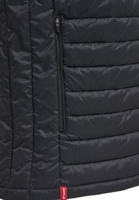 Marine gewatteerde jas met horizontaal doorgestikte panelen, een ritszak aan de zijkant en een rode merketiket bij de onderkant.