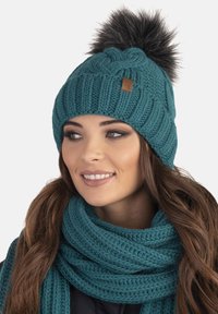 Un bonnet en tricot bleu canard avec un design texturé et un pompon en fausse fourrure sur le dessus, associé à une écharpe assortie en matériau et couleur similaires.