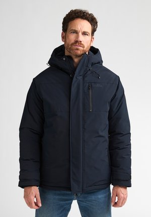 Man with curly hair wearing a black hooded winter jacket and blue jeans standing against a plain white background.