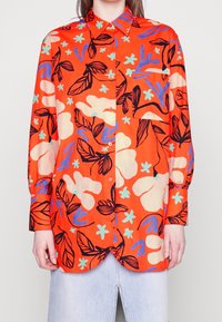 Chemise à boutons en tissu orange vif avec un motif floral noir, bleu et vert menthe ; comprend une poche poitrine et des manches longues.