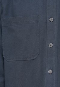 Ύφασμα πουκαμίσου σκούρου μπλε με τσέπη στο στήθος στην αριστερή πλευρά και τρία ορατά γκρι κουμπιά στο μπροστινό μέρος.