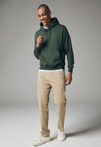 Sudadera de color verde oscuro con un bolsillo frontal y cordones, combinada con pantalones beige y zapatillas blancas. El material parece suave y casual.