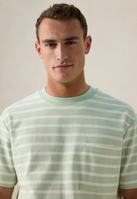 T-shirt rayé vert clair avec des rayures blanches horizontales, col rond et une petite poche poitrine. Tissu doux avec une coupe décontractée.