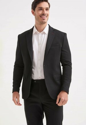 Homme souriant portant une veste de costume noire, une chemise blanche boutonnée et un pantalon noir, debout devant un fond clair uni.