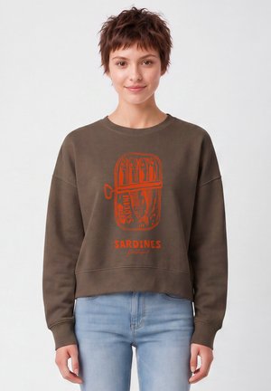 Jeune personne aux cheveux courts et bruns portant un sweat-shirt marron avec un motif rouge de boîte de sardines et un jean bleu clair sur fond blanc.