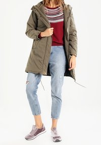 Femme portant un parka vert olive, un pull rouge à motifs, un jean bleu clair déchiré et des baskets mauves, debout devant un fond blanc.