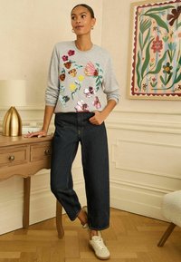 Szara bluza z kolorowymi kwiatowymi zdobieniami z cekinów, zestawiona z ciemnymi dżinsami o szerokich nogawkach i białymi sportowymi butami, na tle jasnego wnętrza.