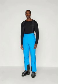 Pantaloni de schi albaștri cu bretele reglabile, buzunare laterale cu fermoar și accente negre la tivuri. Modelul poartă un top negru cu mânecă lungă.