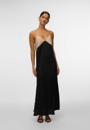 Femme debout portant une robe longue noire et beige sans manches avec des bretelles fines et des sandales noires à bouts ouverts sur un fond clair uni.