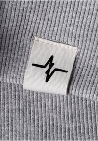 Weißes Stoffetikett mit einer schwarzen Zickzack-Herzschlaglinie, die auf ribbeligem grauen Strickstoff genäht ist.