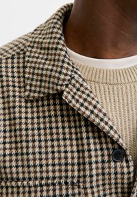 Bruin en beige houndstooth patroon jack met een omklapbare kraag, gecombineerd met een gebreide beige trui met ronde hals. Zwarte knoopdetails.