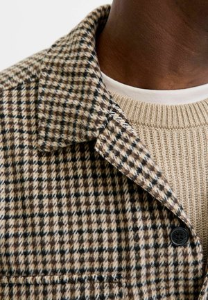 Bruin en beige houndstooth patroon jack met een omklapbare kraag, gecombineerd met een gebreide beige trui met ronde hals. Zwarte knoopdetails.