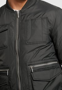 Svart bomberjacka i quiltad nylon, med frontdragkedja, två bröstfickor och två fickor med dragkedja på sidan. Strukturerad tygfinish.