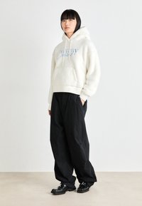 White fleece hoodie with large front pocket and blue logo, paired with loose black trousers and black shoes on a light background.