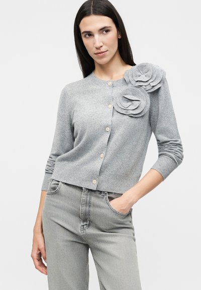 Γυναίκα που φοράει ανοιχτό γκρι ζακέτα με κουμπιά, διακοσμημένη με δύο μεγάλες υφασμάτινες εφαρμογές λουλουδιών στον αριστερό ώμο και τζιν παντελόνι σε ταιριαστό γκρι χρώμα.