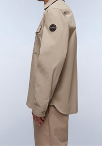 Camicia beige a maniche lunghe con vestibilità rilassata, tessuto morbido, una tasca sul petto e una toppa circolare con logo sulla spalla sinistra.