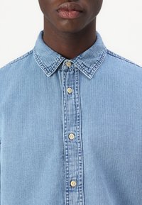 Cămașă din denim de un albastru deschis, cu guler ascuțit, deschidere cu nasturi în față, având nasturi de o culoare deschisă și o textură verticală subtilă.