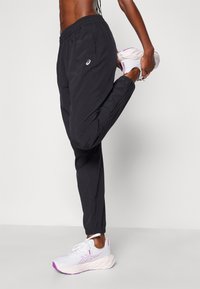 Svarta atletiska joggerbyxor med elastisk midja och siddragkedjor, tillsammans med vita sneakers med lila detaljer.