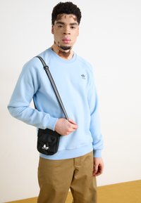 Svetlo modra majica s krožnim izrezom, rebrastimi manšetami in majhnim temnim logotipom. V kombinaciji s khaki hlačami in črno torbico čez telo.