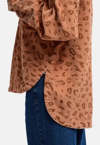 Camicia in corduroy color ruggine con macchie di leopardo marroni, dotata di maniche lunghe, orlo curvo e spacco laterale, abbinata a jeans scuri.
