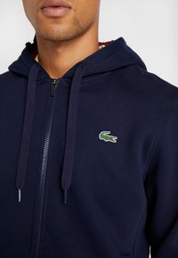 Merimies sininen vetoketjullinen huppari pehmeää kangasta, jossa vasemmassa rinnassa vihreä krokotiililogolla ja teksturoidulla nyörityshupulla.