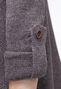 Manica di un indumento grigio lavorato a maglia con texture a coste e dettaglio in bottone marrone sul polsino, che mette in evidenza un orlo arrotolato e un tessuto morbido.