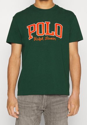 Osoba nosząca ciemnozieloną koszulkę z "POLO Ralph Lauren" dużymi pomarańczowymi literami na piersi oraz jasnoszare spodnie.