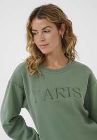 Sweatshirt vert avec l'inscription « PARIS » en lettres en relief. Texture douce, coupe décontractée avec épaules tombantes et encolure ronde.