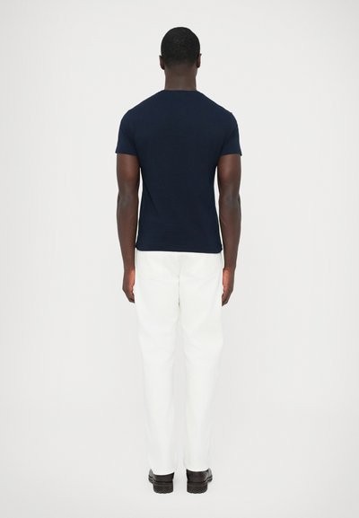 Teget plava majica kratkih rukava s okruglim izrezom, uparena s bijelim hlačama. Glatka tekstura i uski kroj vidljivi s leđa.