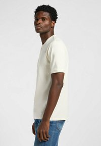 Cremefarvet kortærmet polo skjorte med knaplukning, lavet af glat bomuld. Båret med blå jeans, hvilket viser en tætsiddende silhuet.